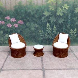 May include: Miniature patio set with two wicker chairs and a small round table. The chairs have white cushions and are made of brown wicker. The table is also brown wicker with a white top. The set is placed on a stone patio.