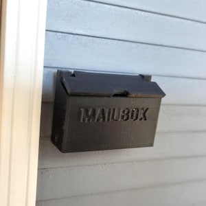 May include: A black mailbox with the word "MAILBOX" embossed on the front is mounted on a light gray, horizontal-planked wall. The mailbox has a hinged lid and is positioned near a white trim and a wooden door frame.