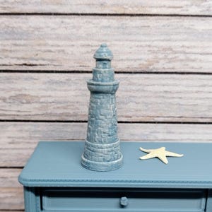 May include: A light blue miniature lighthouse with a textured surface stands on a matching blue table. A small, cream-colored starfish rests on the table's surface. The background features a light wood-grain pattern.