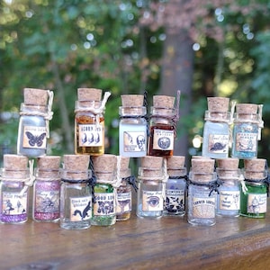May include: A collection of 16 small glass bottles with cork stoppers and twine. Each bottle contains a different colored liquid and has a label with a whimsical Halloween-themed design and text. The labels include text such as "Moth Dust", "Acorn Oil", "Skeleton Elixir", "Snail Slime", "Mermaid Scales", "Pixie Pocket", "Fairy Dust", "Black Cats Whiskers", "Good Luck", "Happy Feet", "Flowers of Belladonna", "Crow's String", "Feathers of Cherub Wings", and "Goblin Water".