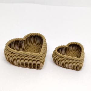 May include: Two brown heart-shaped woven baskets. The larger basket is in the foreground and the smaller basket is in the background.