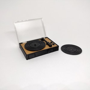 May include: A miniature turntable with a black base and a tan top. The turntable has a black record on the platter and a separate black record. A clear plastic cover is open. The design is simple and modern.