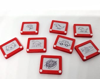 May include: A collection of small, red, toy drawing boards with white screens. Each board features a different black and white drawing, including a flower, dinosaur, cat, house, car, dog, airplane, and scribbles.