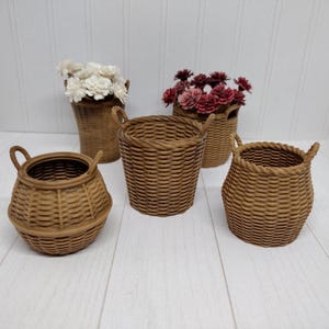 May include: Five small, woven brown baskets of varying shapes and sizes. Two baskets contain white and burgundy flowers. The baskets have handles and are arranged on a white wooden surface, creating a rustic, decorative display.