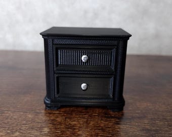 May include: A small, black nightstand with two drawers and silver knobs. The nightstand has a classic design with decorative trim around the edges and on the drawer fronts. The nightstand is sitting on a wooden surface.