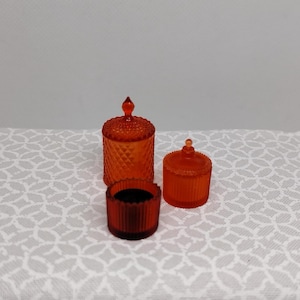 May include: Three orange glass containers with lids. The tallest container has a diamond pattern, while the smaller one has vertical lines. The smallest container is a dark orange color. The containers are on a white patterned surface.