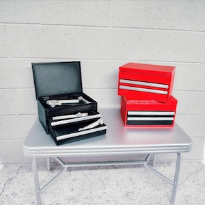 May include: A black textured tool chest with an open lid and three drawers containing small silver wrenches, positioned next to a stack of two red tool chests with silver drawer handles. The items are arranged on a silver-colored table surface.