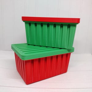 May include: Two rectangular storage containers, one stacked on top of the other. The bottom container is red with a green lid, and the top container is green with a red lid. Both have vertical ribbed sides.