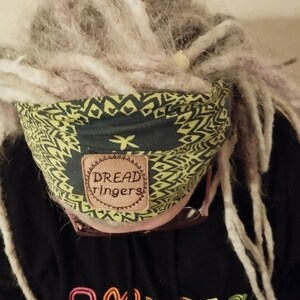 May include: A person with long white dreadlocks is wearing a green and yellow patterned headband with a brown leather patch that says "Dread ringers".