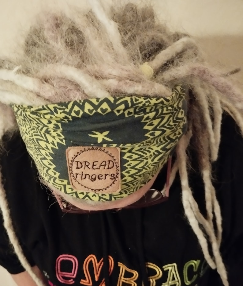 May include: A person with long white dreadlocks is wearing a green and yellow patterned headband with a brown leather patch that says "Dread ringers".