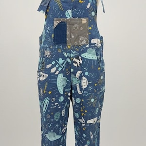 May include: Blue denim dungarees with a space-themed pattern of rockets, planets, and UFOs. They have tie straps and a front pocket with a patch. The garment is displayed on a mannequin.