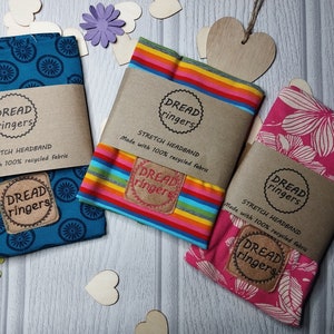 May include: Three colorful stretch headbands made with 100% recycled fabric. The headbands feature a blue and white pattern, a rainbow stripe pattern, and a pink floral pattern. Each headband has a brown label with the text "Dread ringers" and "Stretch Headband" printed on it.