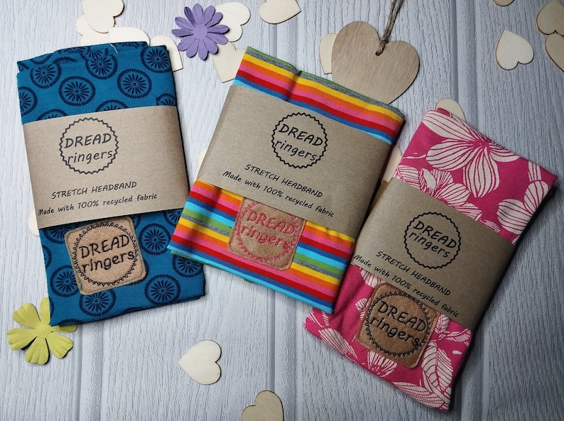 May include: Three colorful stretch headbands made with 100% recycled fabric. The headbands feature a blue and white pattern, a rainbow stripe pattern, and a pink floral pattern. Each headband has a brown label with the text "Dread ringers" and "Stretch Headband" printed on it.