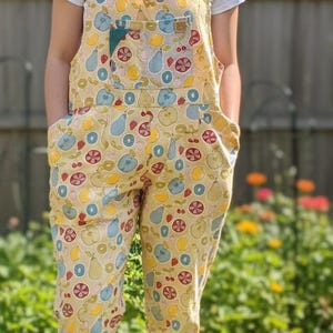 May include: Yellow overalls patterned with fruit, including apples, pears, and strawberries in red, blue, and yellow. The dungarees have a bib pocket and side pockets. A white t-shirt is worn underneath.