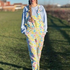 May include: Light-coloured overalls featuring a playful pattern of cartoon characters in pastel shades. The person is wearing a light blue sweatshirt. The overalls have a bib and pockets.