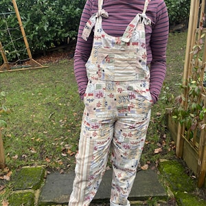 May include: Beige patchwork overalls with red and blue floral and geometric patterns. The dungarees have a front pocket and tied shoulder straps. A striped long-sleeved top is worn underneath. The person is wearing black shoes.