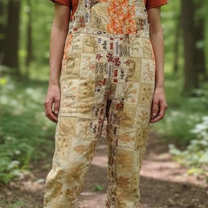 May include: A person wearing tan patchwork overalls with a rust-coloured t-shirt. The overalls feature a variety of patterned squares in beige, orange, and red. Brown hiking boots with red laces complete the outfit. The background is a blurred forest.