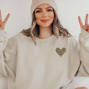 Può includere: Felpa beige con un design a cuore leopardato. La felpa ha girocollo e maniche lunghe. È visibile anche un berretto beige.