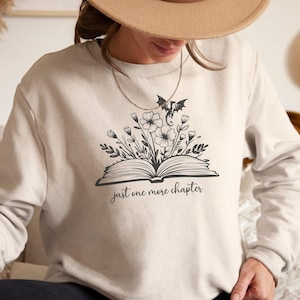 Könnte beinhalten: Beiges Sweatshirt mit einer schwarzen Grafik eines offenen Buches mit Blumen und einer Fledermaus. Der Text "just one more chapter" steht unter dem Buch. Das Sweatshirt hat lange Ärmel.