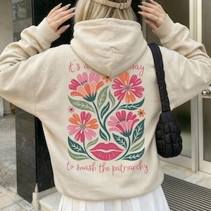 Könnte beinhalten: Beige Kapuzenpullover mit floralem Design auf dem Rücken, mit rosa, orangefarbenen und grünen Blumen und dem Text "It's a good day to smash the patriarchy". Der Kapuzenpullover hat eine Kapuze mit Kordelzug und lange Ärmel.