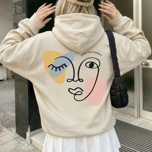 Könnte beinhalten: Ein beigefarbener Kapuzenpullover mit einer schwarzen Strichzeichnung eines Gesichts auf der Rückseite, ergänzt durch gelbe und rosa abstrakte Formen. Der Text "SMASH THE PATRIARCHY" steht über dem Gesicht. Kombiniert mit einer beigen Kappe.