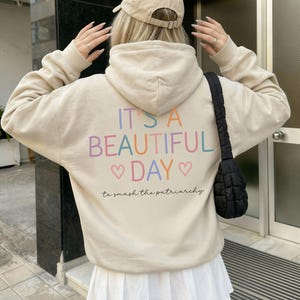 Puede incluir: Una sudadera con capucha beige con el texto "IT'S A BEAUTIFUL DAY" en colores pastel, con corazones y la frase "to smash the patriarchy" debajo. La persona lleva una gorra de béisbol beige y una falda plisada blanca.