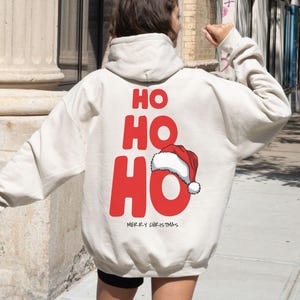 Könnte beinhalten: Ein cremefarbener Kapuzenpullover mit den Worten "HO HO HO" in großen roten Buchstaben auf dem Rücken, zusammen mit einer Weihnachtsmützen-Grafik. Der Satz "MERRY CHRISTMAS" ist darunter gedruckt. Der Hoodie hat eine Kapuze und lange Ärmel.