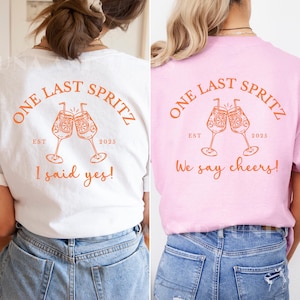 Könnte beinhalten: Zwei T-Shirts, eines weiß und eines rosa, mit dem orangefarbenen Schriftzug "ONE LAST SPRITZ" und zwei Cocktailgläsern. Das Design beinhaltet "EST 2025". Das weiße Shirt sagt "I said yes!" und das rosa sagt "We say cheers!"