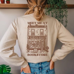 Puede incluir: Sudadera beige con un gráfico marrón de una librería y el texto "MEET ME AT BOOKSTORE". El gráfico de la librería incluye el texto "Booked & Bound". La sudadera la lleva una persona con pelo rubio.