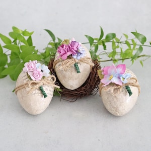 May include: Three decorative egg-shaped ornaments. Each egg is wrapped with paper and twine, and topped with small, colorful flowers. One egg rests in a small nest. Green foliage is in the background.