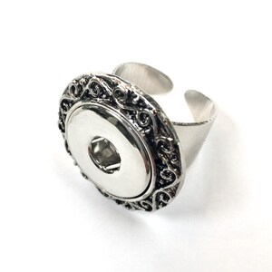May include: A silver adjustable ring with a decorative filigree design around a snap-on button. The ring is designed to hold a 18mm snap-in charm or design.