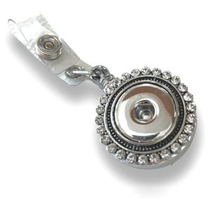 May include: A silver retractable badge reel with a clear plastic strap and a decorative, round face. The face is embellished with a ring of clear rhinestones around a silver button. The badge reel is designed to hold an ID badge.