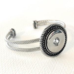 May include: A silver bracelet with a double-strand cable design and a round snap button charm. The charm has a black and silver design.