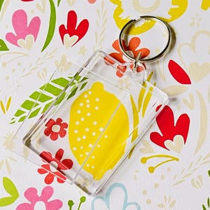 May include: Clear rectangular keychain with a yellow lemon design and a silver key ring. The lemon has white dots and is set against a floral background with red, pink, and green accents. A fun and colorful accessory.