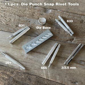 May include: A set of 11 silver metal tools for making snap rivets. The tools include a die base with different size holes, a punch tool, and a setting tool. The tools are laid out on a wooden surface.