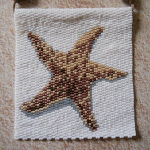 May include: A white beaded wall hanging featuring a brown starfish design. The hanging has a brown leather strap with two fish-shaped charms.