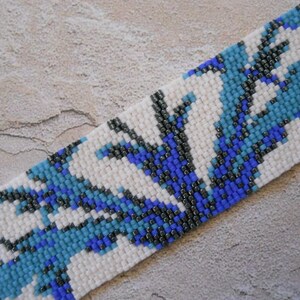 May include: A beaded bracelet with a white background and a pattern of blue and black beads. The pattern resembles stylized flowers or leaves.