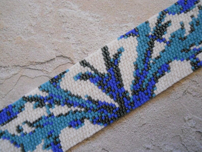 May include: A beaded bracelet with a white background and a pattern of blue and black beads. The pattern resembles stylized flowers or leaves.