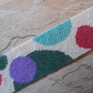 May include: A beaded bracelet with a white background and colorful geometric shapes. The shapes are in shades of blue, green, purple, and pink.