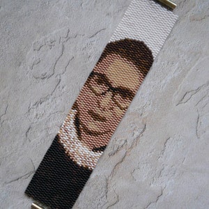 May include: A beaded bracelet with a portrait of Ruth Bader Ginsburg. The bracelet is made with black, brown, white, and pink beads. The bracelet has gold metal clasps.