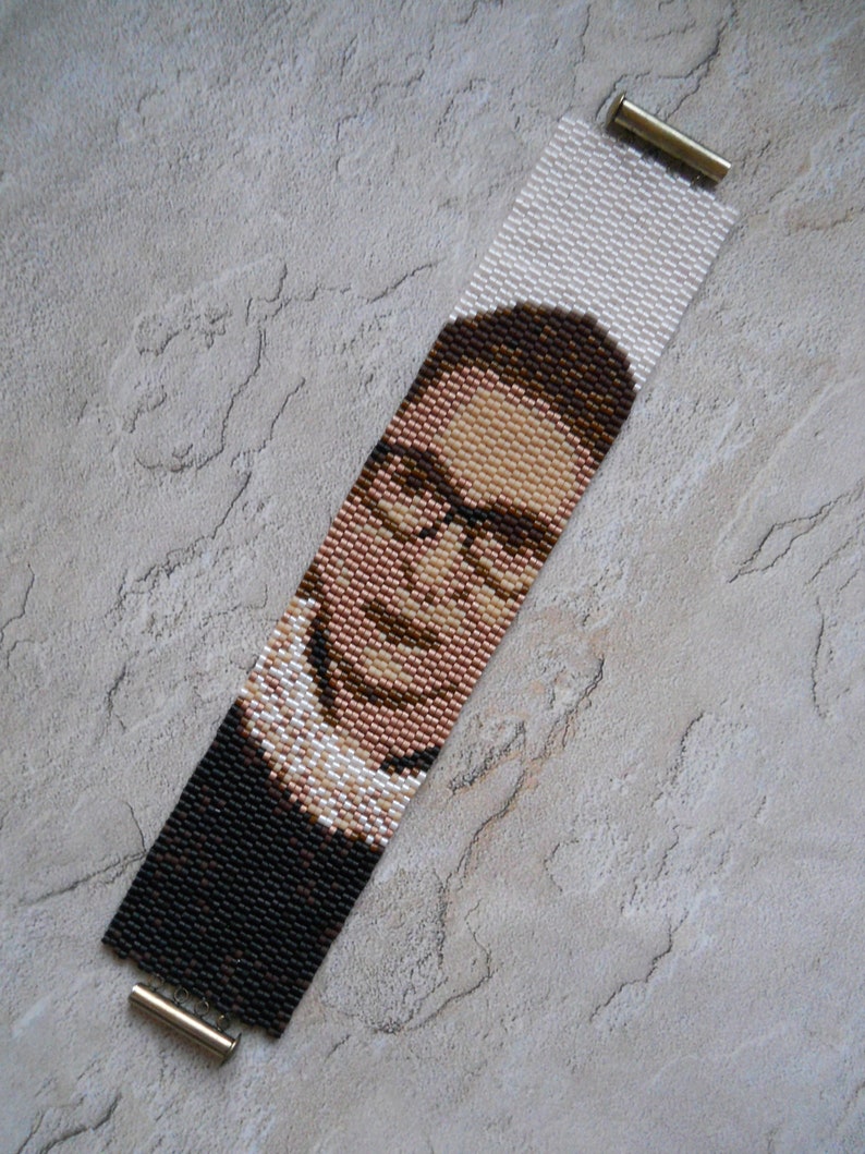 May include: A beaded bracelet with a portrait of Ruth Bader Ginsburg. The bracelet is made with black, brown, white, and pink beads. The bracelet has gold metal clasps.