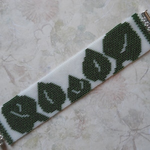 Op de afbeelding: Een witte kralen armband met een patroon van groene bladeren. De armband heeft zilveren sluitingen aan beide uiteinden.