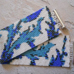 May include: A white beaded bracelet with a blue and black geometric pattern. The bracelet has silver metal clasps.
