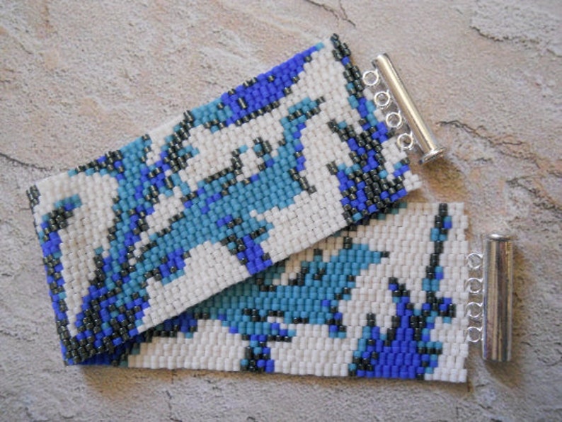 May include: A white beaded bracelet with a blue and black geometric pattern. The bracelet has silver metal clasps.
