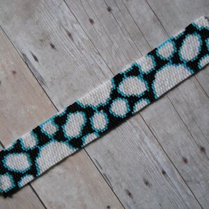 May include: A white, black, and turquoise beaded bracelet with a geometric pattern. The bracelet has silver clasps on each end.