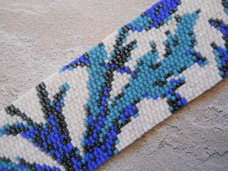 May include: A close-up of a beaded bracelet with a white background and a pattern of blue, black, and gray beads.