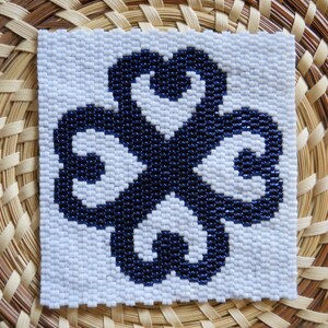 May include: A white square with a blue four-leaf clover design. The clover is made of small, dark blue beads.
