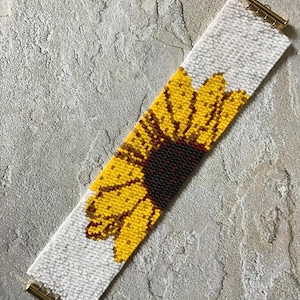 May include: A white beaded bracelet with a yellow sunflower design. The bracelet has gold metal clasps.