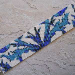 May include: A white beaded bracelet with a blue and black geometric pattern. The bracelet is made with small seed beads and has a silver clasp.
