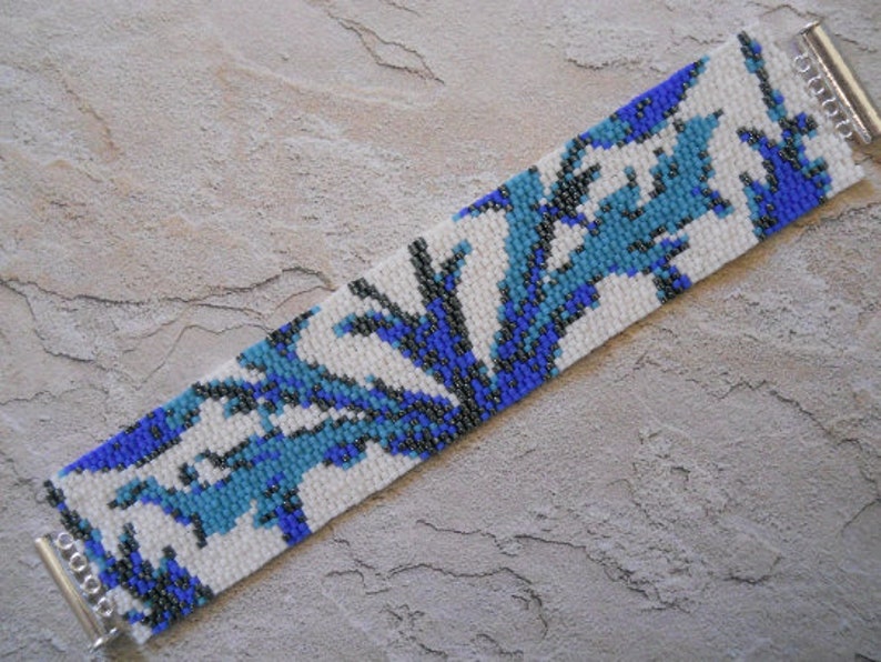 May include: A white beaded bracelet with a blue and black geometric pattern. The bracelet is made with small seed beads and has a silver clasp.